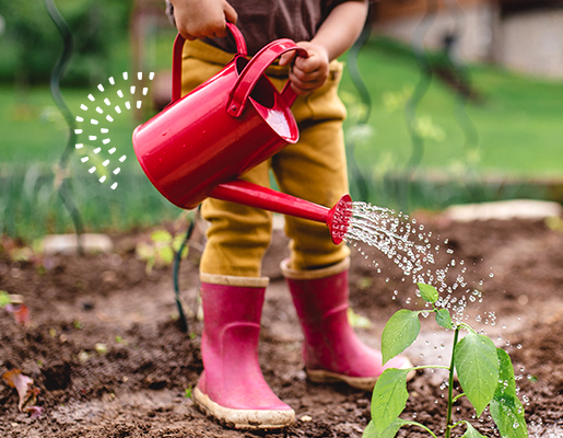 A kid wearing red boots, watering a plant with a red watering can.