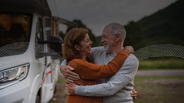 Two people hugging near a recreational vehicle with hills and trees in the background.