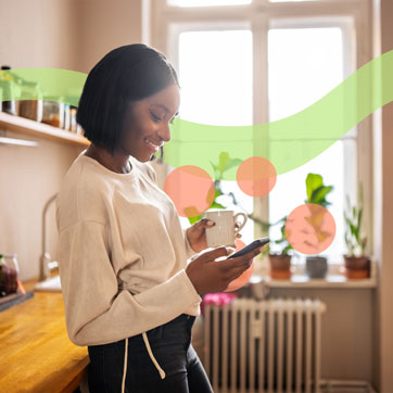 A woman standing in her kitchen holding a cup of coffee and her cellphone.