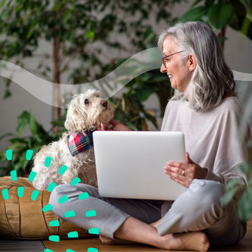 A woman with gray hair pets her dog while she has a laptop in her lap.