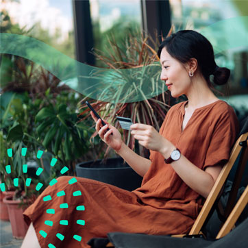 A woman is sitting outside holding a credit card up with her left hand and her cell phone with her right like she's typing in her card information.