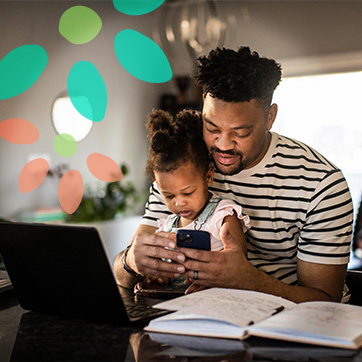 A man holds a child on their lap while they both look at a smartphone. They are seated at a table with an open laptop, notebook, and papers.