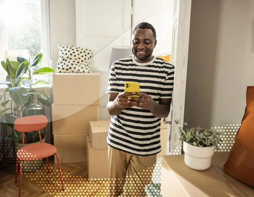 Indoor scene of a person holding a phone, surrounded by packed boxes, chairs, and a potted plant.