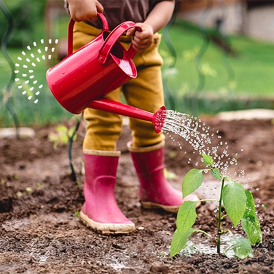 Child wearing red boots waters a small plant with a red watering can in a garden bed.