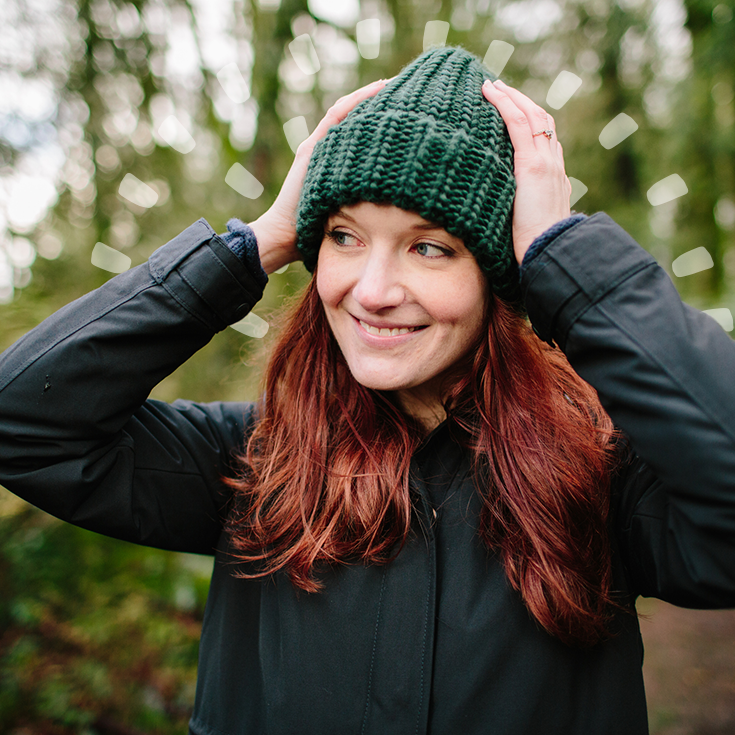 Person in a dark coat holding a teal knitted hat on their head while standing on a forest path.
