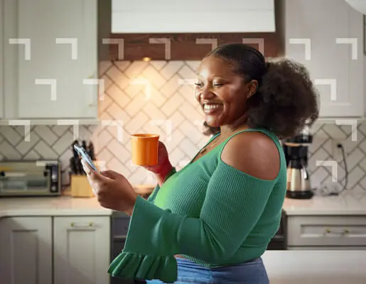 A person holding a mug looks at a smartphone while standing in a well-lit kitchen.